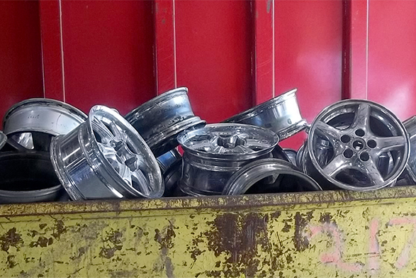 Dynamic Metal Recycling | A collection of discarded metal car rims, destined for industrial recycling, are piled in a worn yellow dumpster against a red background.
