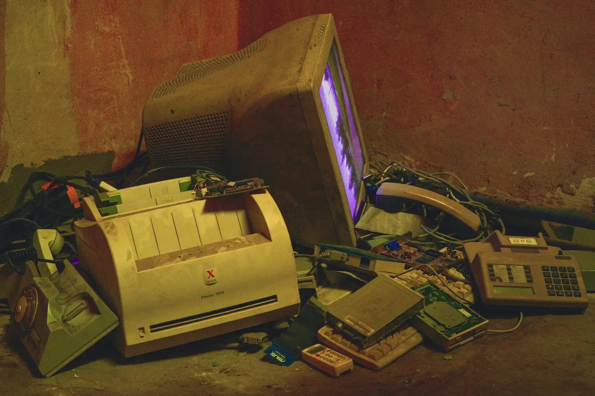 Dynamic Metal Recycling | A dusty CRT monitor, old printer, circuit boards, telephone, and calculator are piled on a dirty floor against a reddish wall.